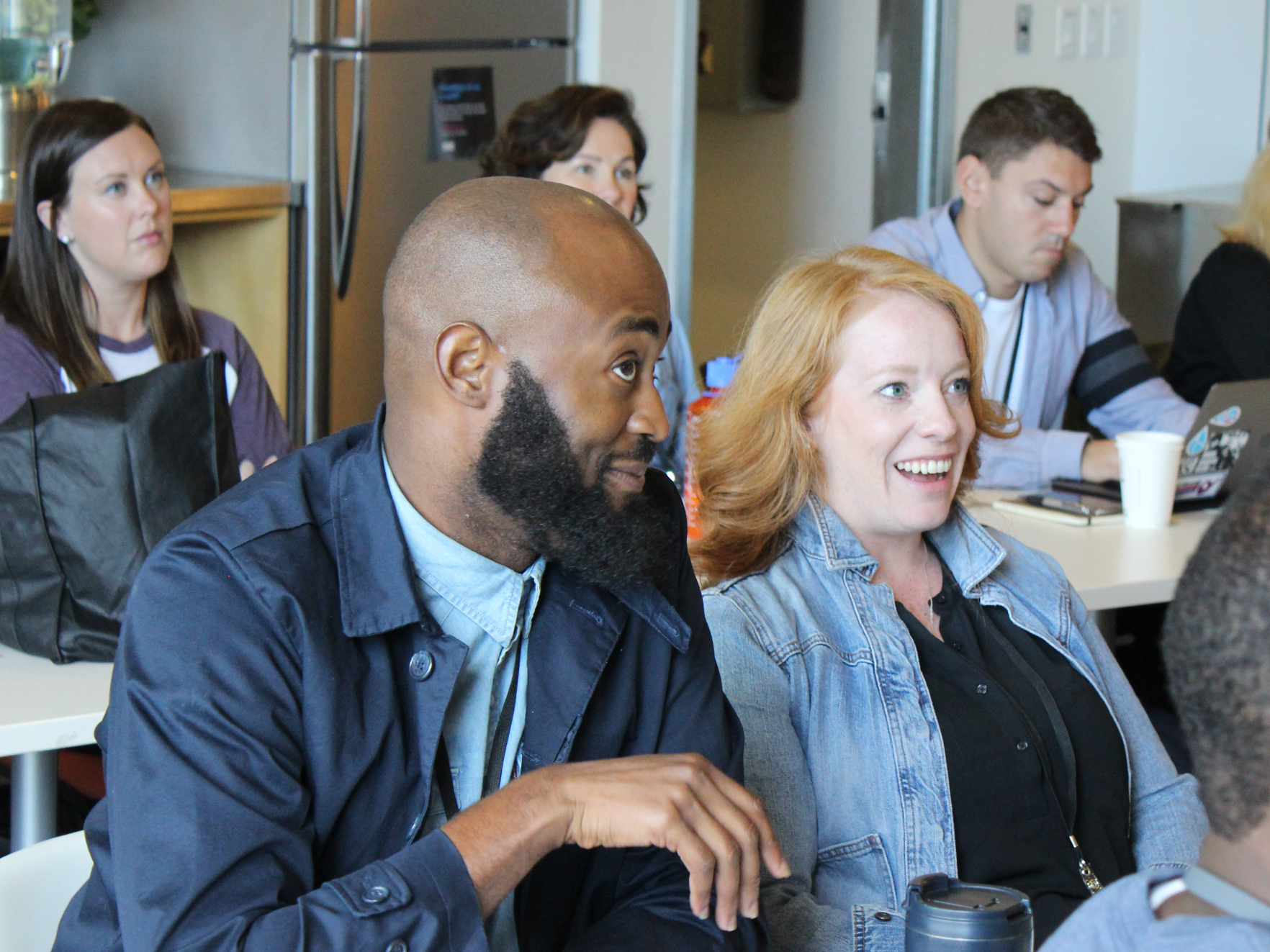 An African American man with a beard and a caucasian woman with red hair sit in a crowd. They are listening for more information.