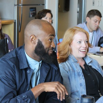 An African American man with a beard and a caucasian woman with red hair sit in a crowd. They are listening for more information.